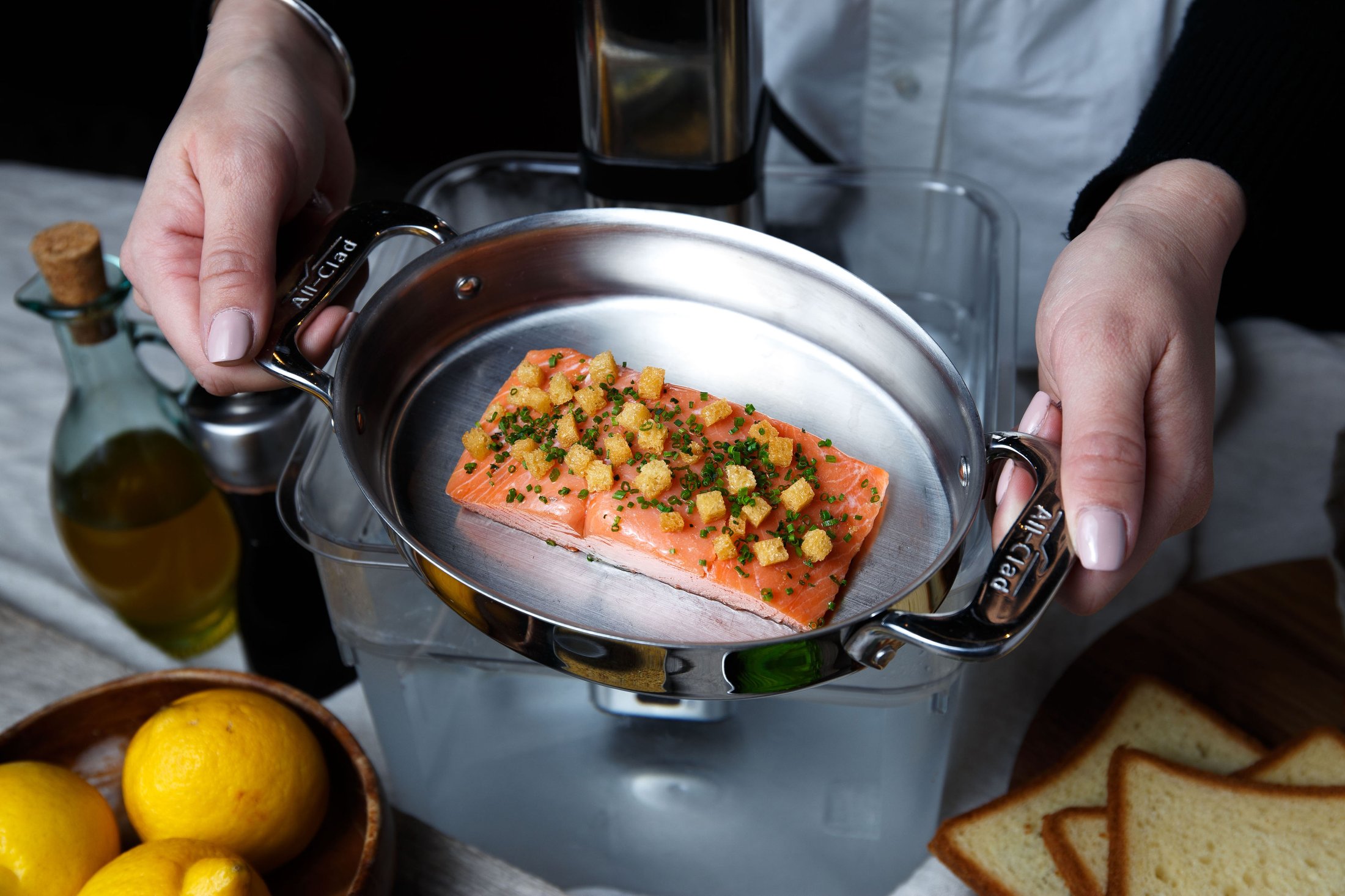 A cured salmon dish with fried croutons and chives being plated in an All-Clad pan